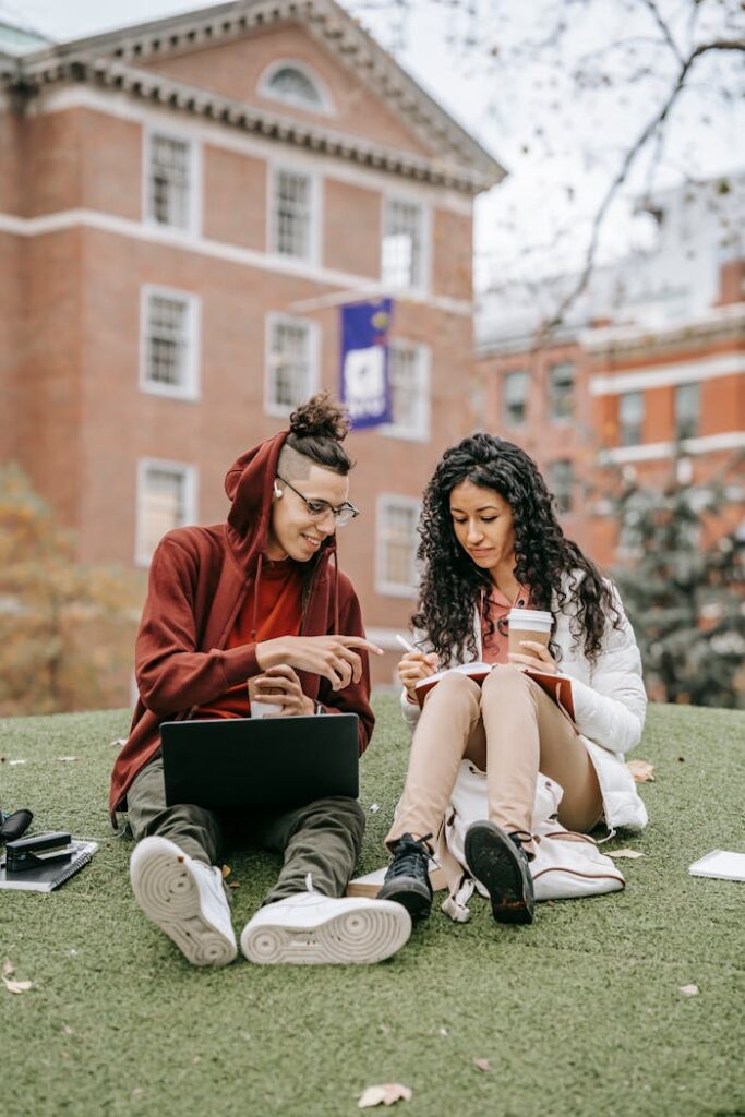 Young multiracial students in outerwear sitting on grass and studying while writing notes in notebook with pen and cup of drink while using netbook in campus in daylight